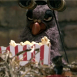 Puppet of a pigeon with an unimpressed look on its face. There is a bowl of popcorn in front and the pigeon is raising a pair of binoculars.