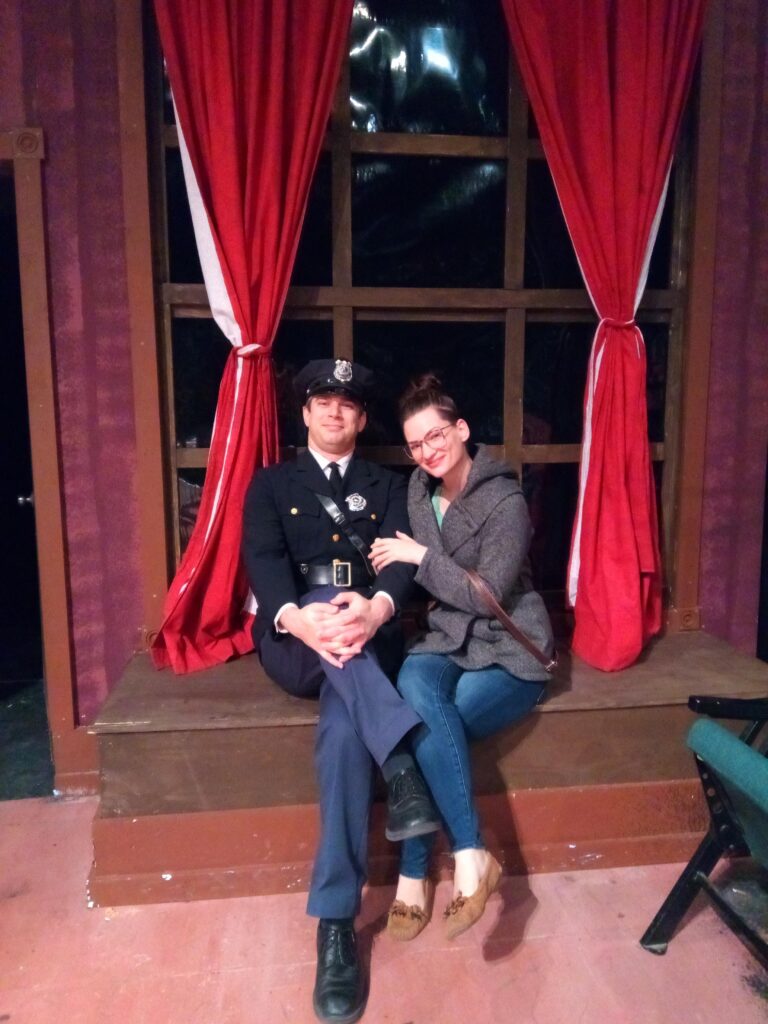 Benton and Abi on the set of the play Arsenic & Old Lace. They are sitting on the bench that the body is hidden in. They are in front of a window. Benton is wearing the police uniform of his character officer O'Hara.