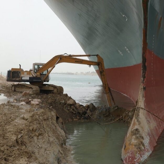 The Ever Given, a ship stuck in a sandbar in the Suez Canal. A backhoe pushing on it from shore looks tiny in comparison.